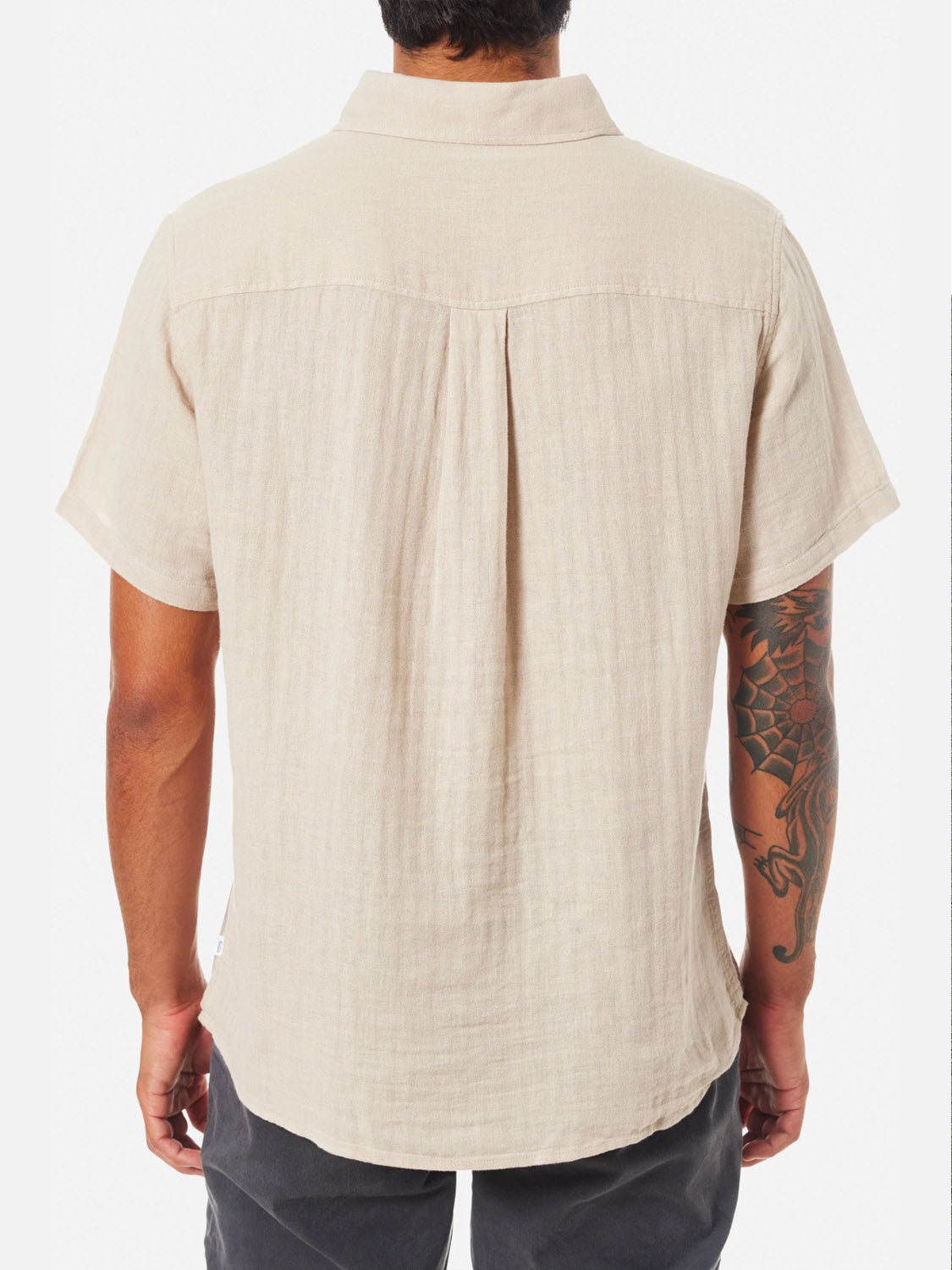 A man faces away, wearing the Katin Alan Solid Shirt in light beige. This short-sleeve button-up by Katin features a relaxed fit and versatile style. His tattoo sleeve is visible against a plain white background.