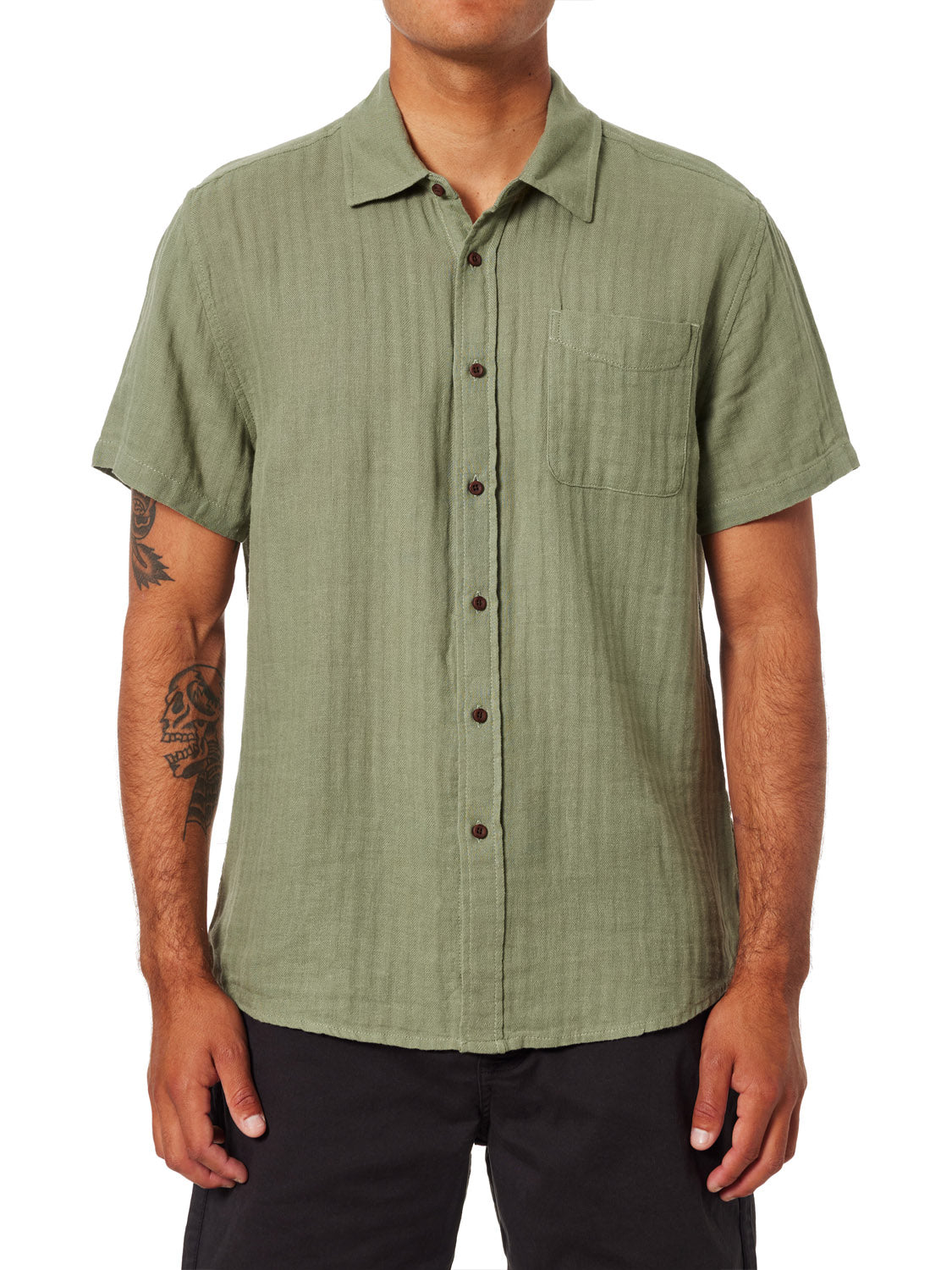 A person wears the Katin Alan Solid Shirt by Katin, a short-sleeve light green button-up with a chest pocket and black pants. Relaxed arms reveal tattoos, highlighting this wardrobe essential against a plain white background.