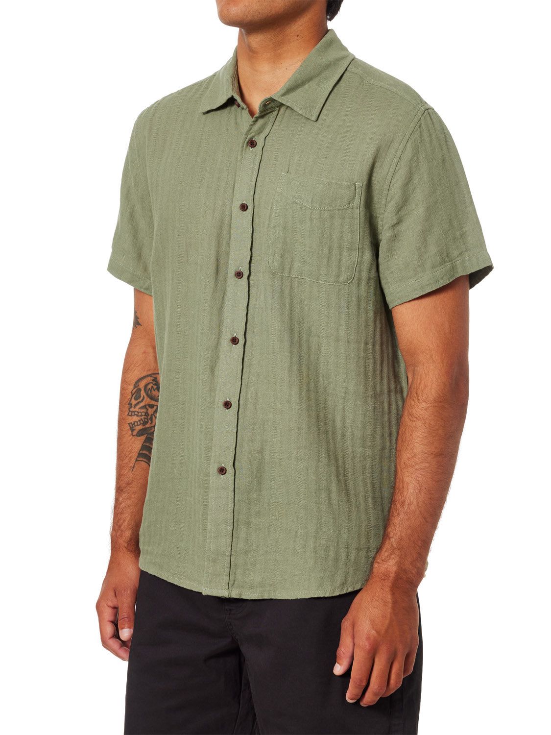 A person wears the Katin Alan Solid Shirt by Katin—a light green, short-sleeve button-up with a chest pocket—paired with black pants. A tattoo is visible on their left forearm. Face not visible.