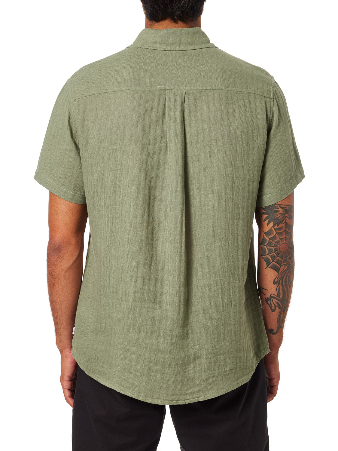A person with a tattoo sleeve is seen from the back wearing the Katin Alan Solid Shirt by Katin in light olive green. This relaxed-fit, short-sleeve button-up features a center pleat and is paired with black pants for versatile style.