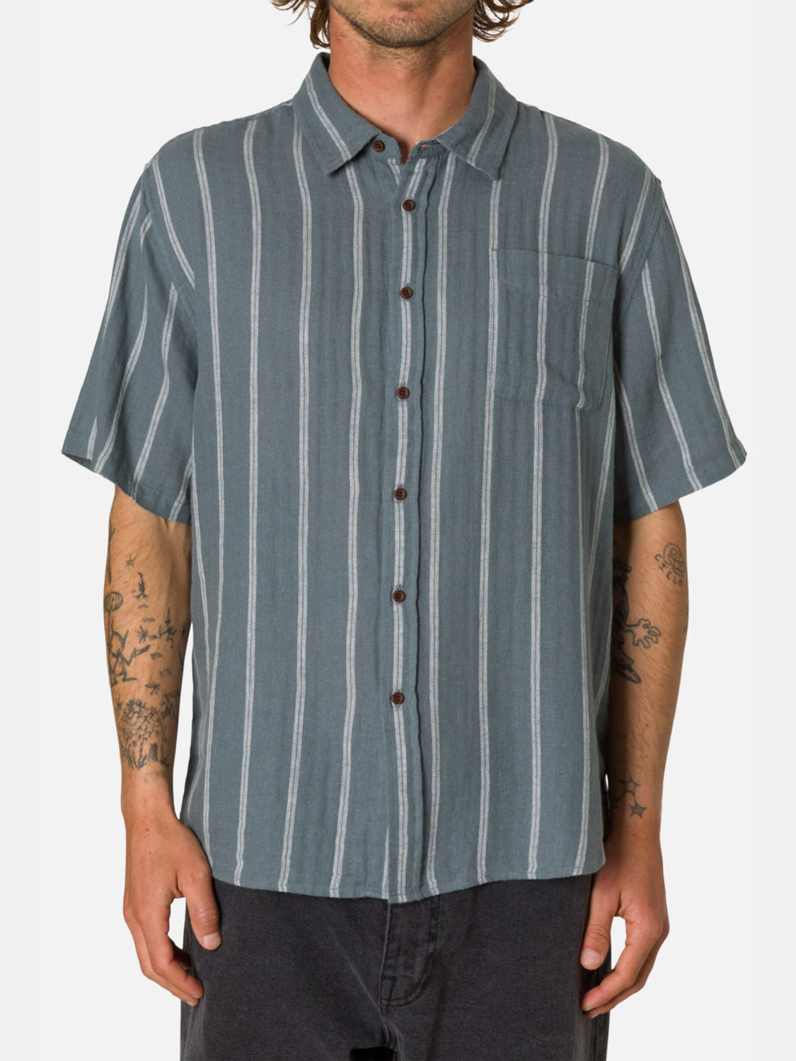A person wears the Katin Alan Short Sleeve Stripe Shirt, featuring blue-gray cotton-linen with vertical white stripes and a chest pocket, paired with dark shorts. Tattoos are visible on both arms.