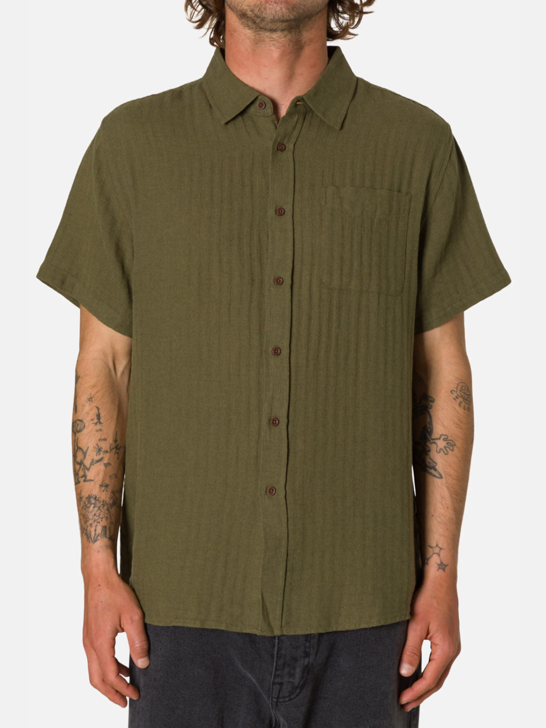 Wearing the Katin Alan Solid Short Sleeve Shirt, a classic olive green cotton-linen shirt with a chest pocket, a tattooed person poses against a plain white background with their face out of view.