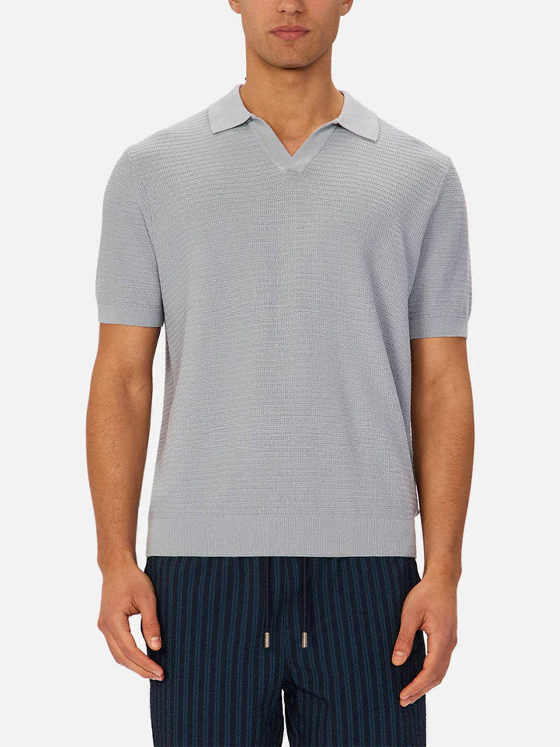 A man stands against a plain white background wearing Industrie’s The Alicante Polo in light gray knitted cotton with an open collar, paired with navy blue striped drawstring pants.