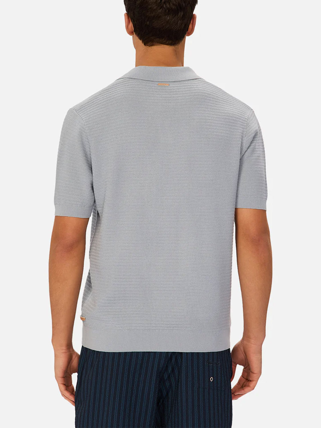 A man is seen from behind wearing Industrie’s The Alicante Polo, a light gray knitted cotton shirt, paired with dark blue pinstriped shorts against a plain white background.
