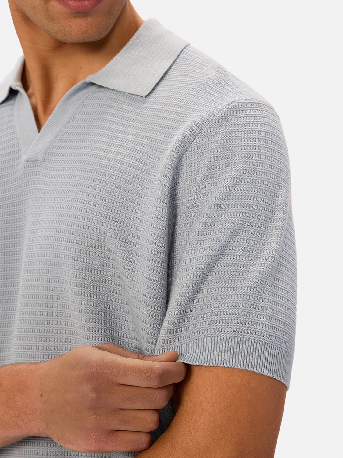 A person adjusts the sleeve of The Alicante Polo by Industrie, a light gray textured knitted cotton shirt. Only the lower face and upper torso are shown against a plain background.