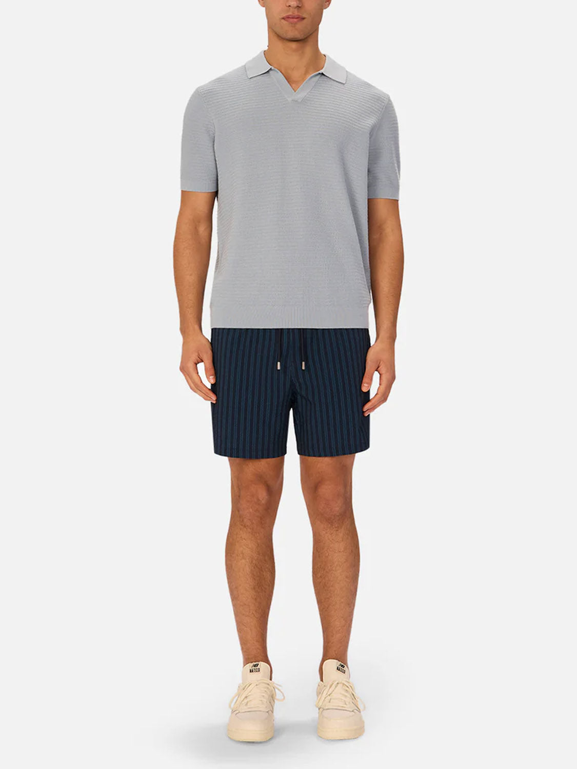 A man wearing Industrie’s The Alicante Polo in light gray, navy blue pinstriped shorts, and cream-colored sneakers stands against a plain white background.