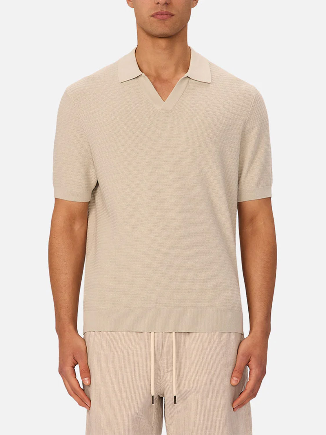 A person wears The Alicante Polo by Industrie, a beige knitted cotton polo shirt with an open collar, paired with beige drawstring shorts, standing against a plain white background.