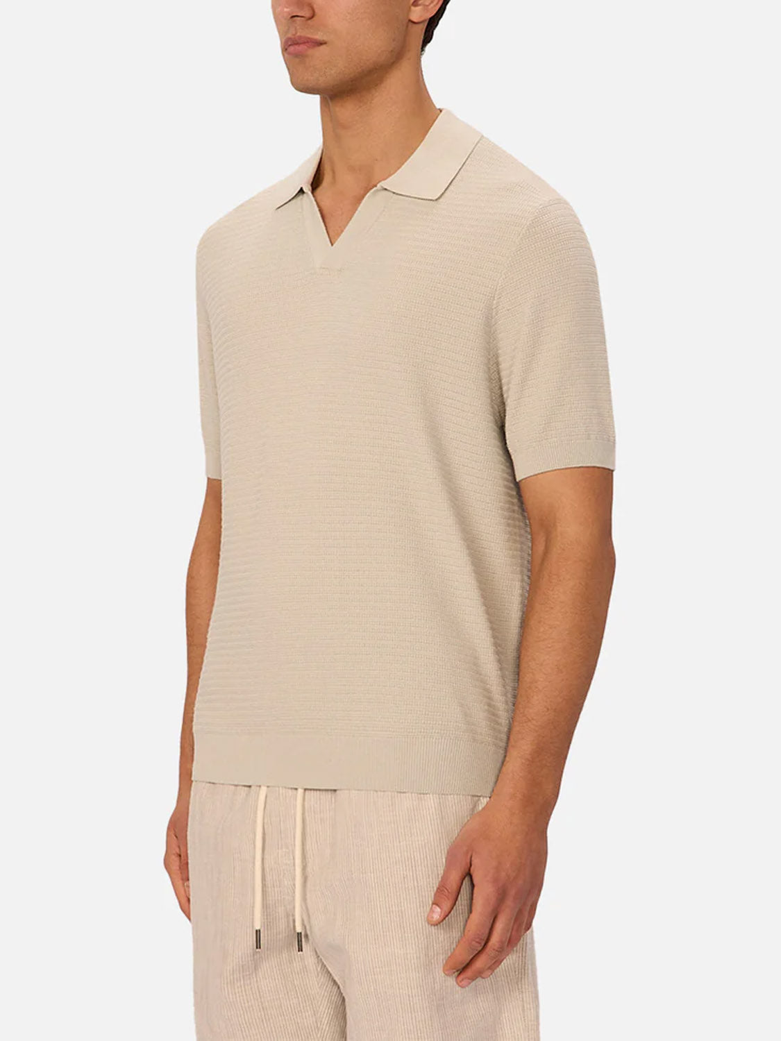 A man wears Industrie’s The Alicante Polo, a light beige textured short-sleeve knit shirt with a split collar and matching drawstring pants, standing against a plain white background.