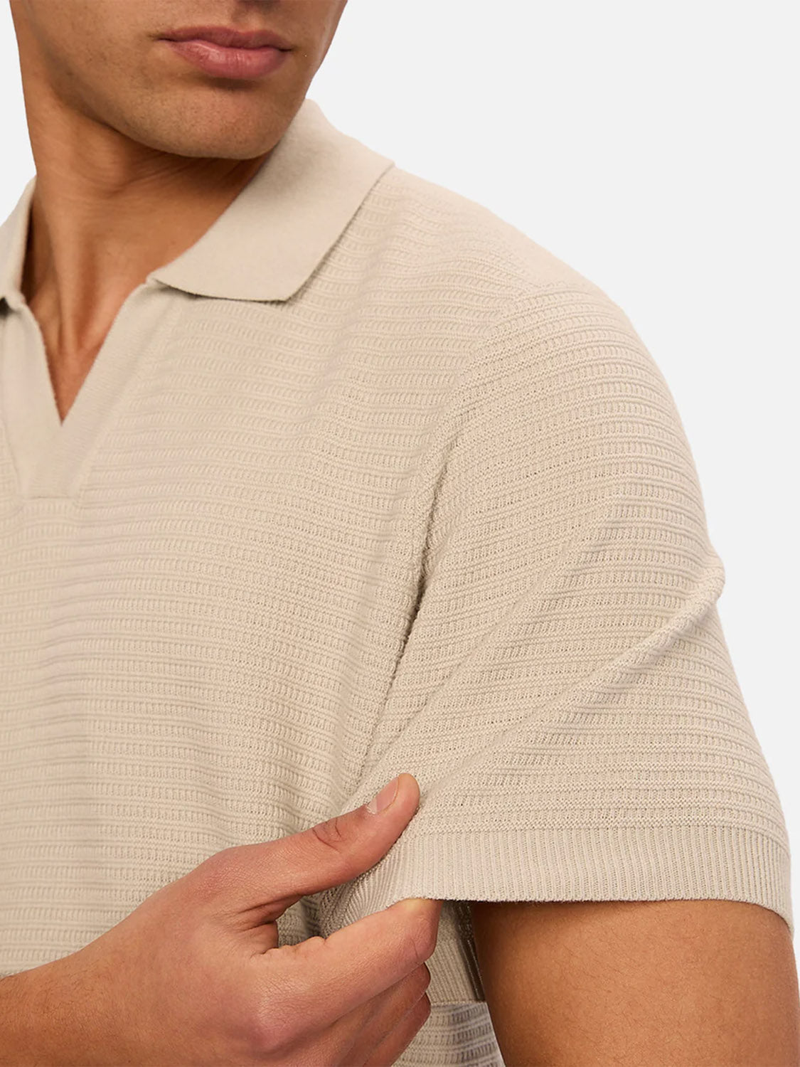 A man in Industrie’s The Alicante Polo in beige subtly tugs at the sleeve, displaying its textured pattern. With only his lower face and upper torso visible, the classic polo’s understated style takes center stage.