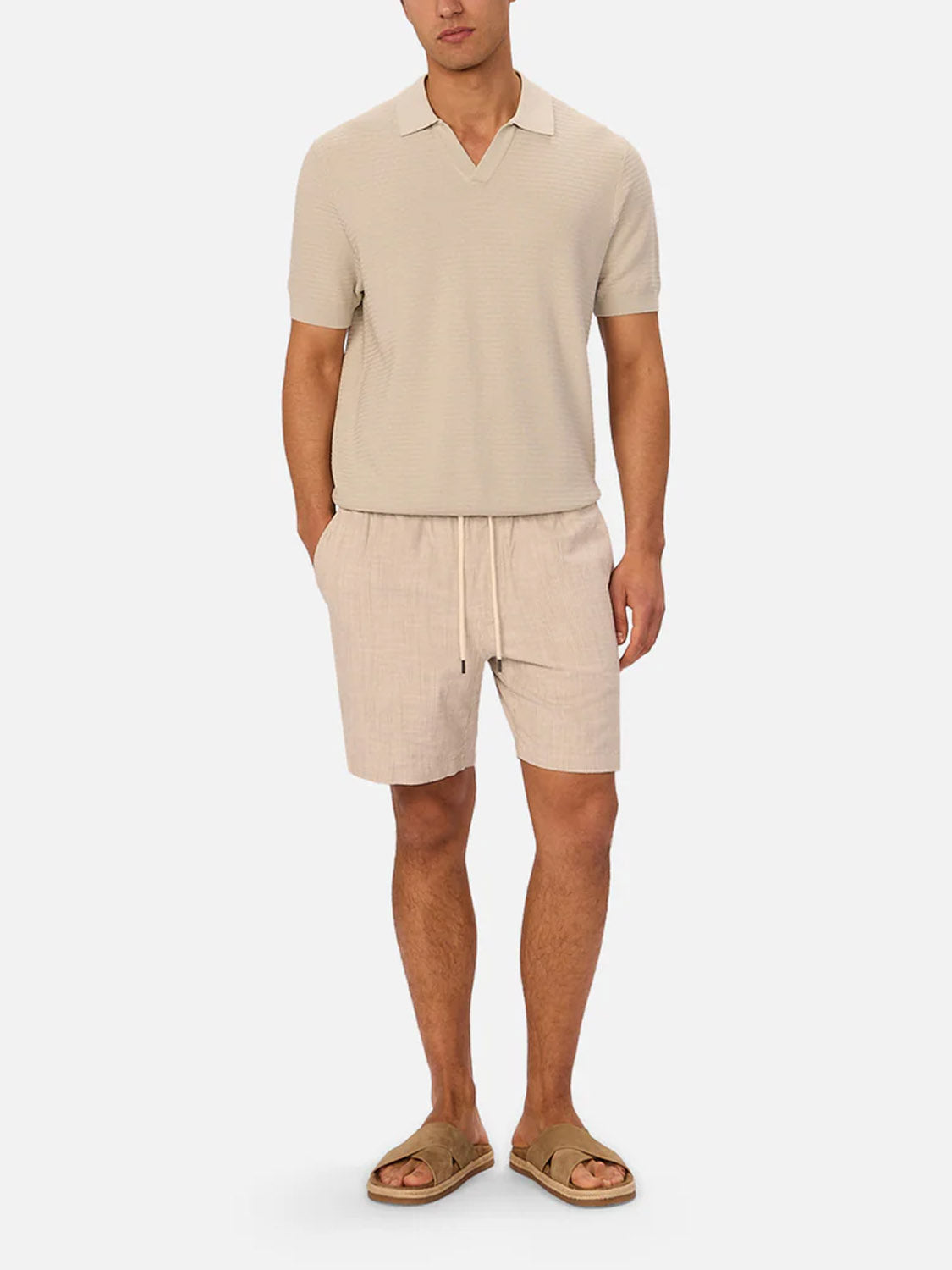 A man wears The Alicante Polo by Industrie in beige, paired with light beige drawstring shorts and tan slip-on sandals, standing against a plain white background with his hands in his pockets.
