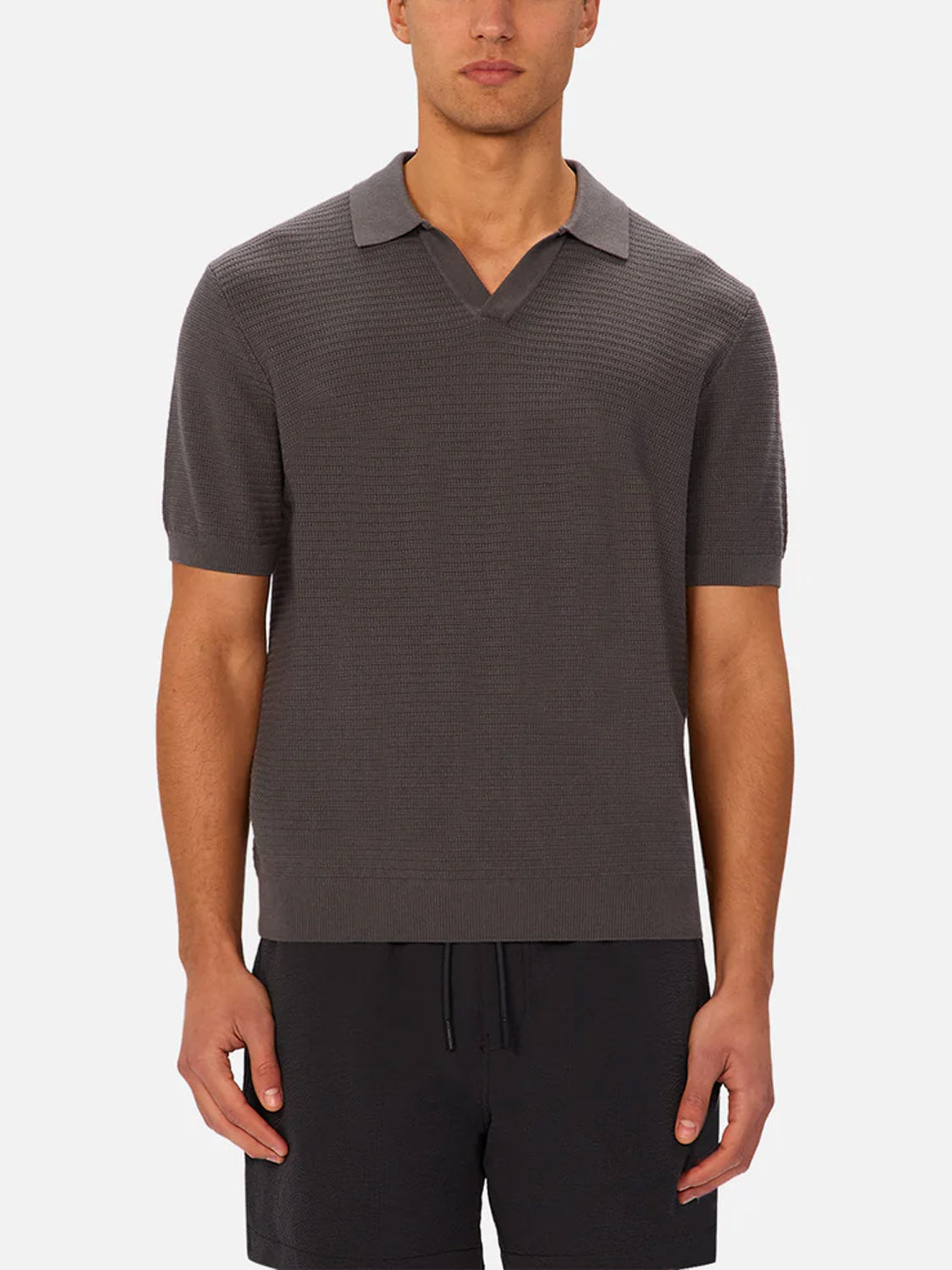 A man wearing Industrie’s The Alicante Polo, a short-sleeve dark gray textured polo with a notch collar, and black drawstring shorts, stands against a plain white background.