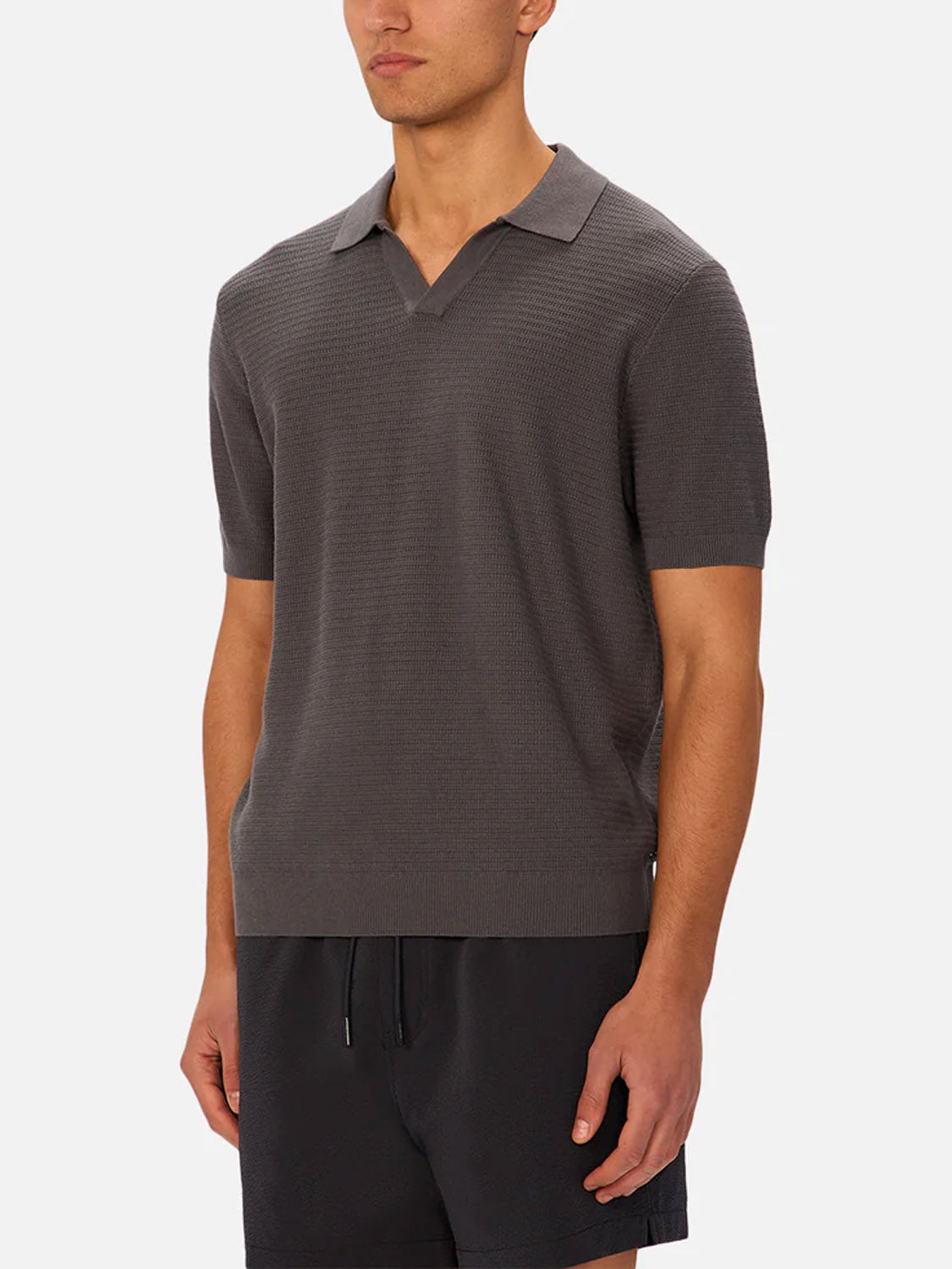 A man wears Industrie’s The Alicante Polo—a short-sleeved, dark gray knitted cotton shirt with a collar and v-neck—paired with black shorts, standing against a plain light background.