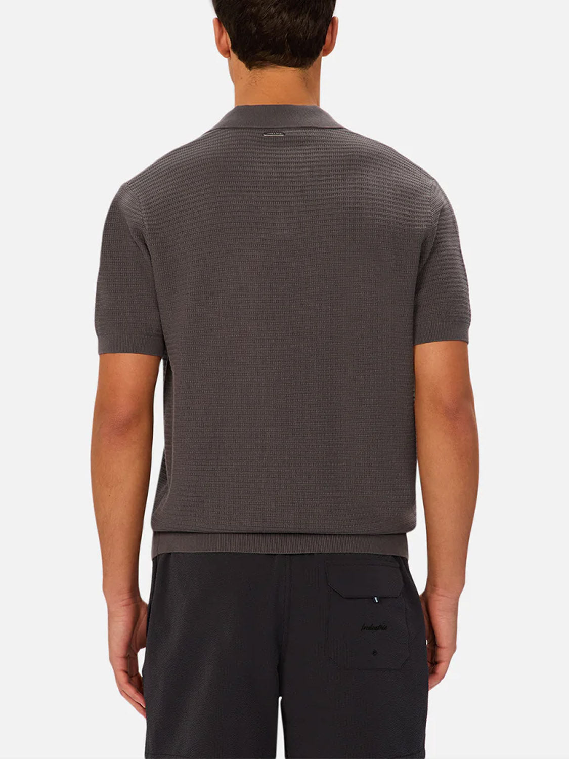 A man is seen from the back wearing The Alicante Polo by Industrie, a short-sleeve dark gray textured shirt, paired with black shorts featuring a back pocket, all set against a plain white background for a refined casual look.