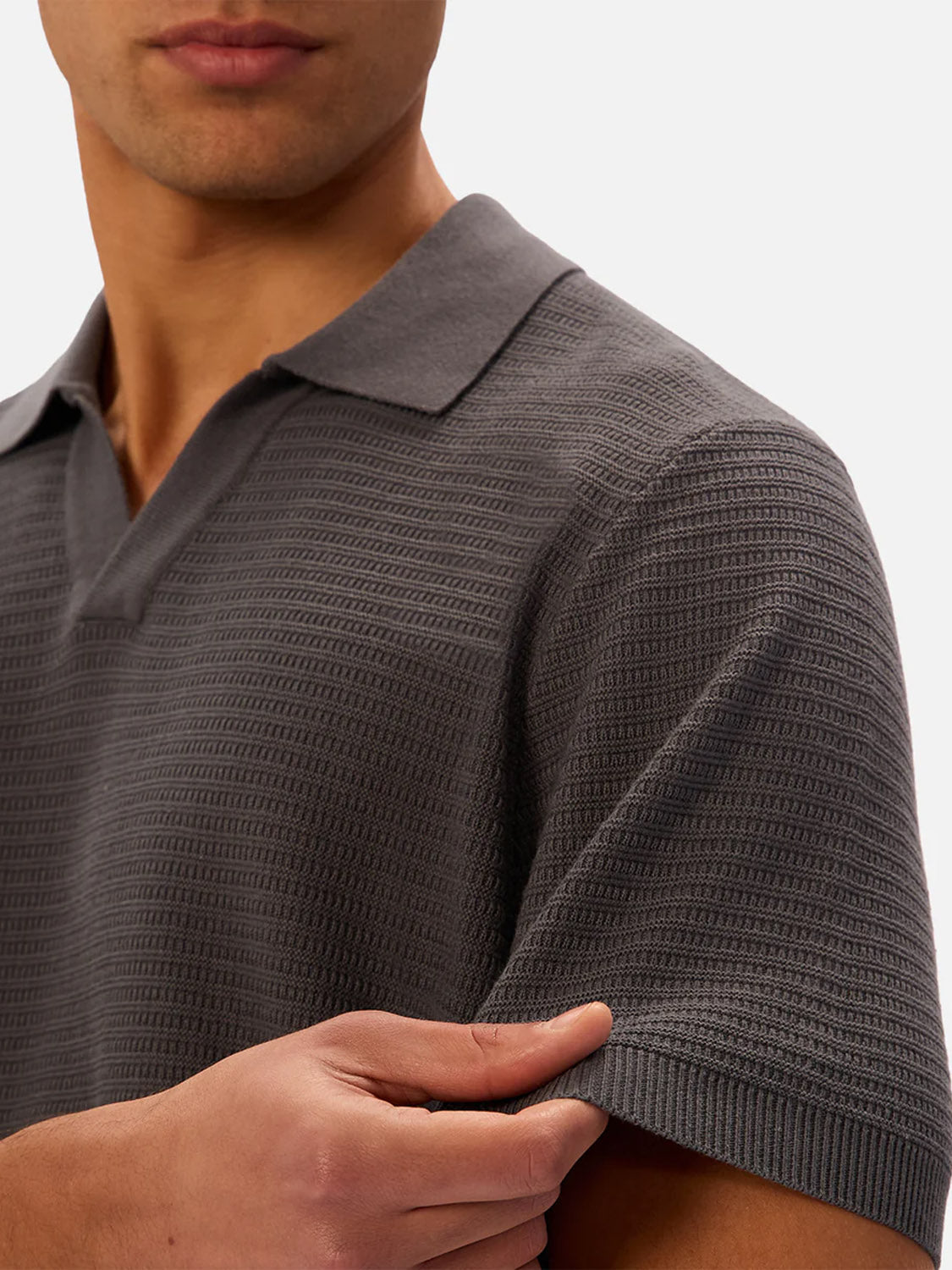 A man stands against a plain background, gently holding his sleeve while wearing The Alicante Polo in charcoal gray by Industrie.