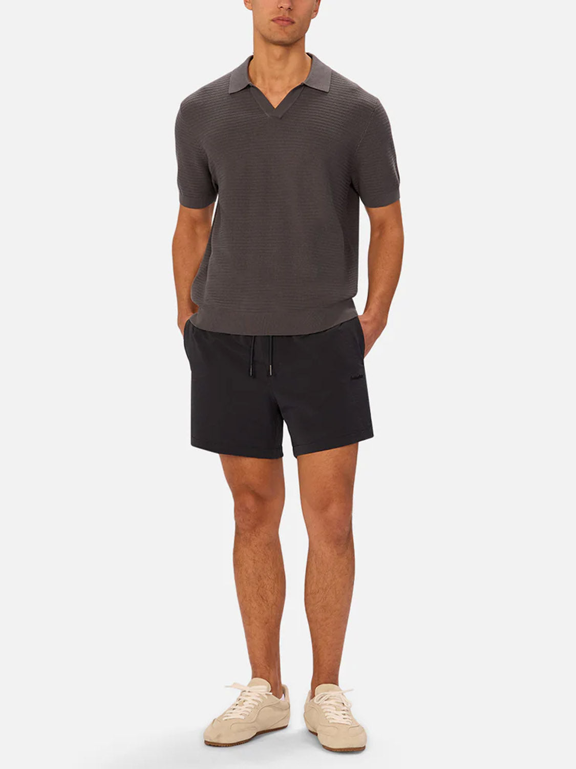 A man stands against a plain background wearing Industrie’s The Alicante Polo, a short-sleeve textured shirt in dark gray, paired with black drawstring shorts and beige sneakers. His hands are in his pockets and he has a neutral expression.