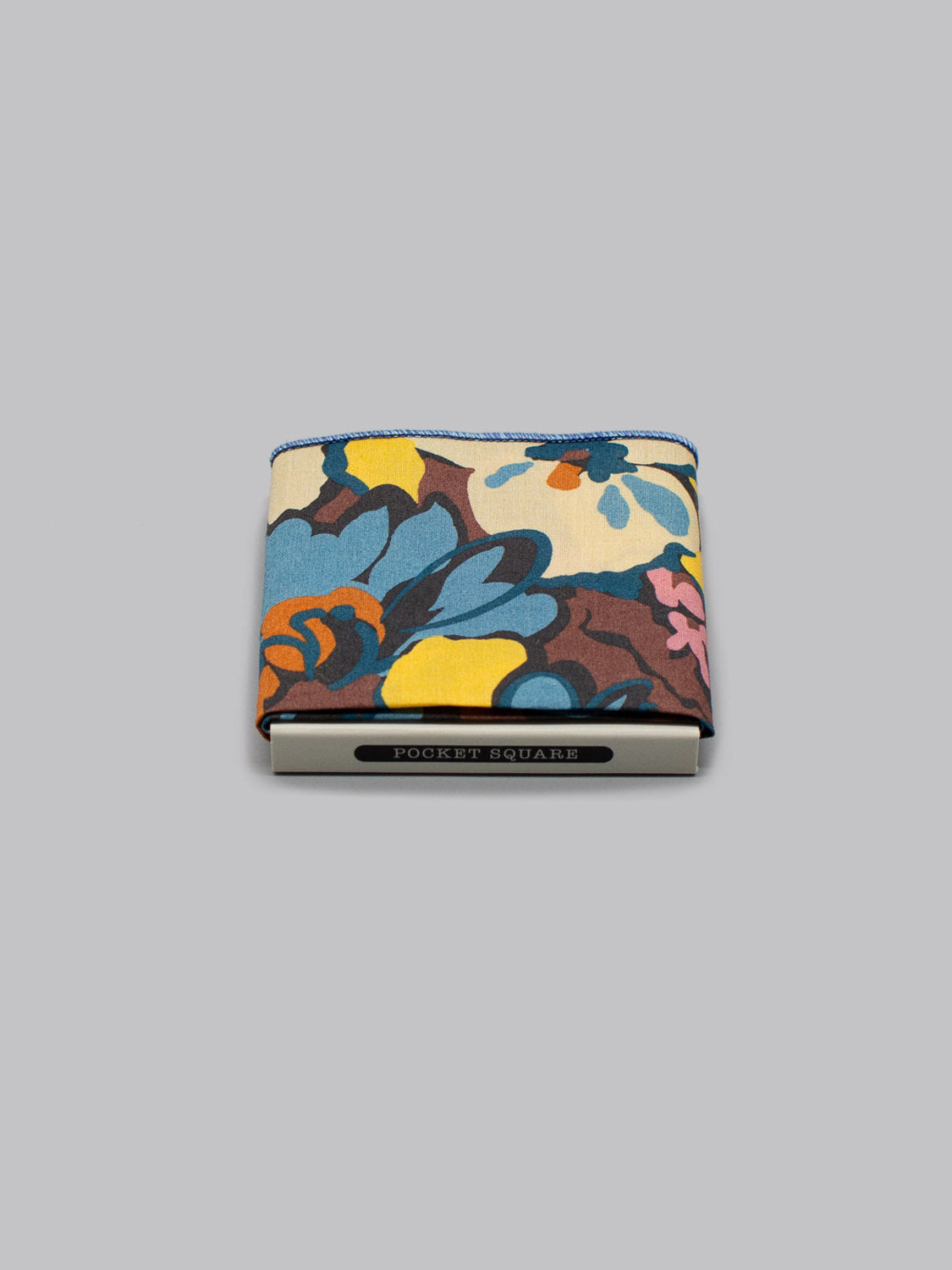 The Liberty™ Alison Pocket Square by ID Menswear, handmade from Liberty of London Tana Lawn with a vibrant yellow, blue, and red floral pattern, is shown folded in a white POCKET SQUARE box on a plain gray background.