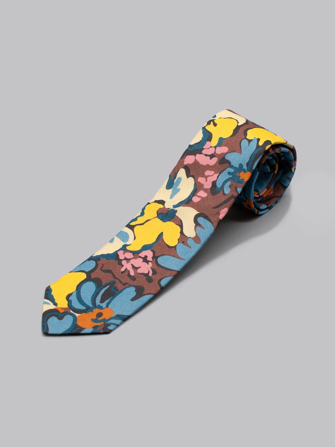 The Liberty of London Tana Lawn™ Alison tie by ID Menswear is handmade in Brooklyn, showcasing a vibrant blue, yellow, pink, and brown floral print on light gray—neatly rolled with the pointed end facing forward.