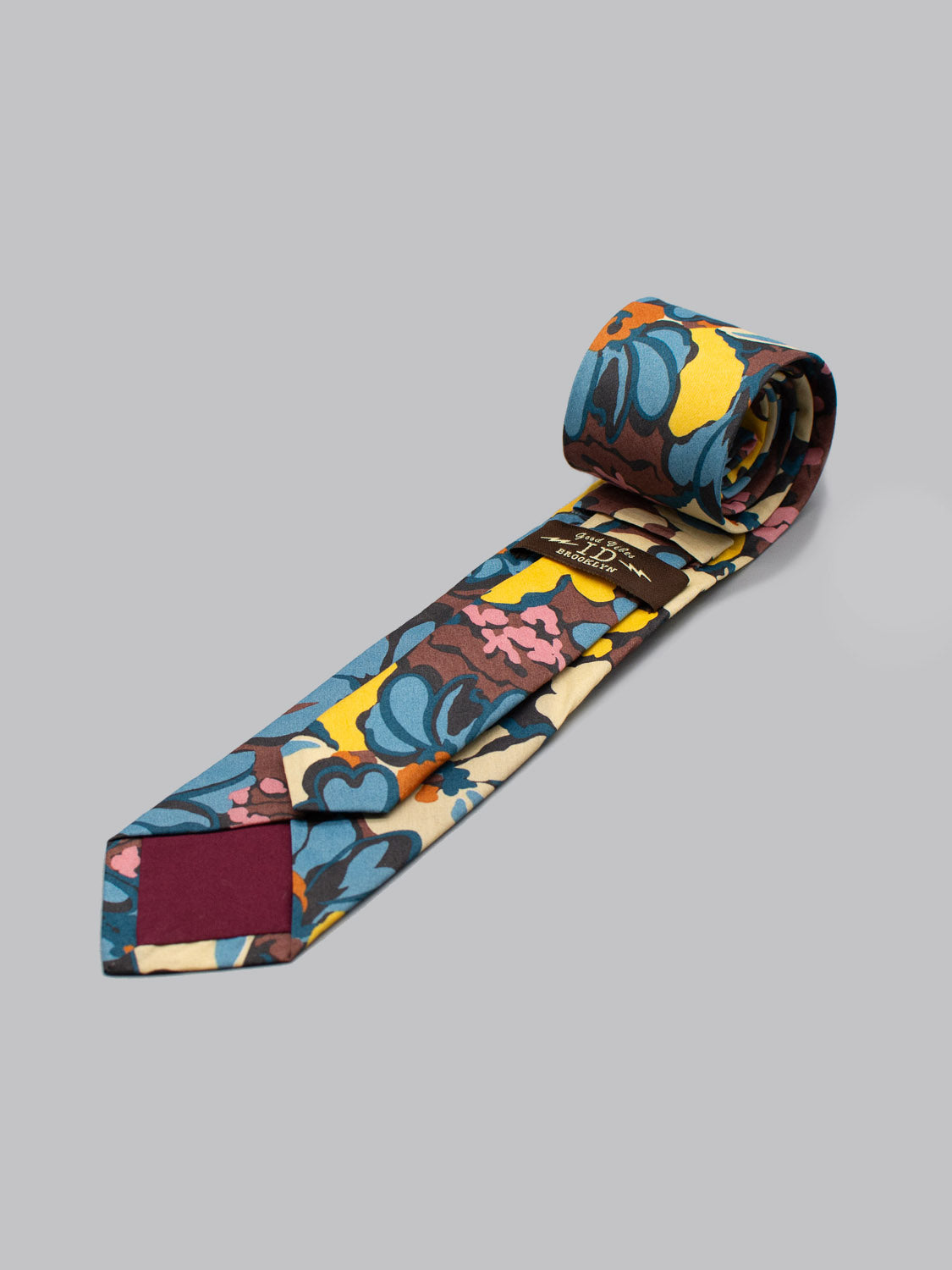 The Liberty of London Tana Lawn™ Alison tie by ID Menswear features a bold abstract floral in blue, yellow, brown, and maroon. Handmade in Brooklyn from luxe cotton, its shown rolled up on a light gray background.