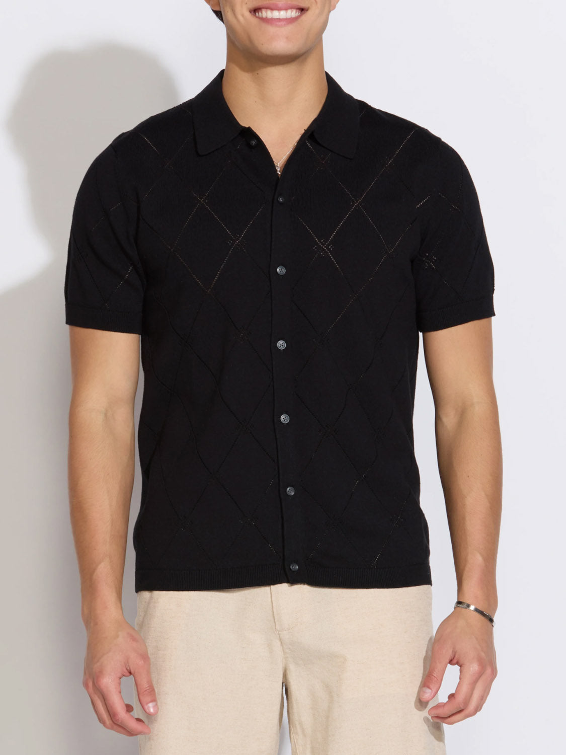 A man smiles wearing the Civil Society Aleppo Full Placket Sweater Polo—a short-sleeve, black top with a subtle diamond pattern—paired with light beige pants.