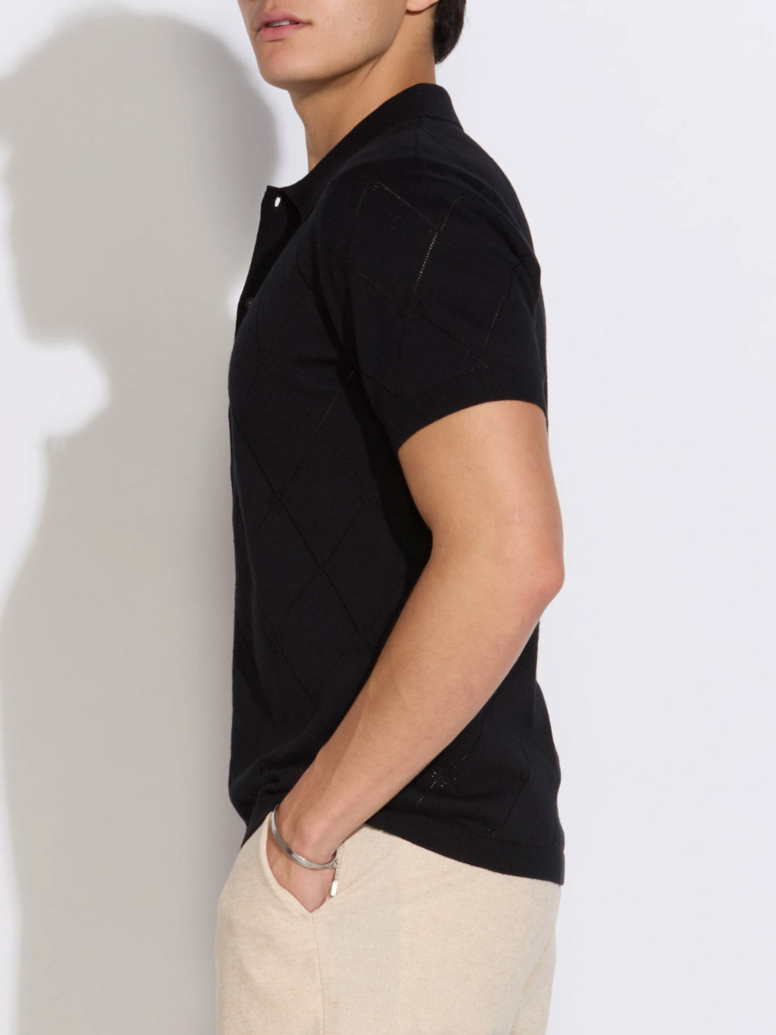 A person stands sideways with one hand in their pocket, wearing the Civil Society Aleppo Full Placket Sweater Polo in black and light beige pants against a plain white background.