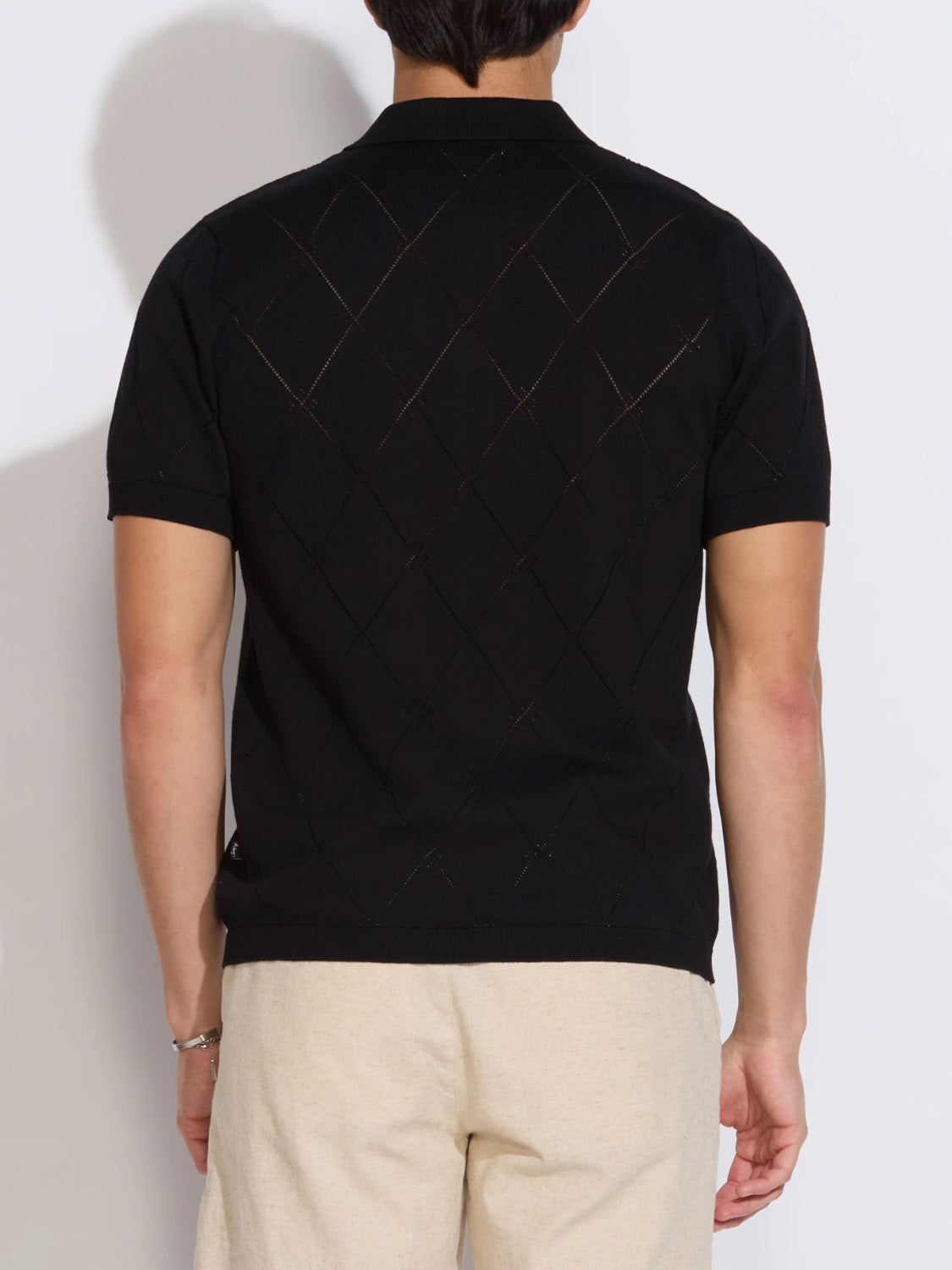 A person wears the Civil Society Aleppo Full Placket Sweater Polo, featuring short sleeves and a diamond pattern on the back, paired with light beige pants. They stand with their back to the camera against a plain white background.