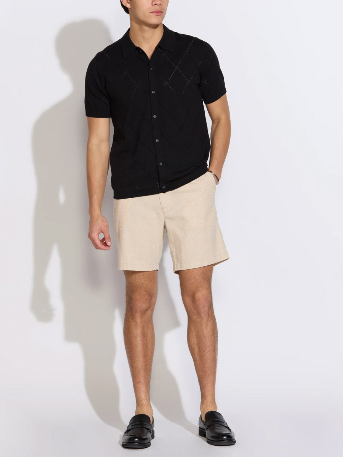 A man stands against a white background wearing Civil Society’s Aleppo Full Placket Sweater Polo in black, paired with beige shorts and black loafers. His face is not shown and his left hand is in his pocket.