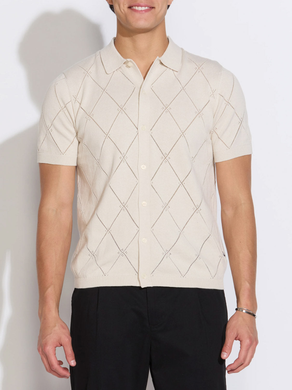 A person wears the Civil Society Aleppo Full Placket Sweater Polo—a cream-colored, diamond-patterned, short-sleeve cotton top—paired with black pants, standing against a plain white background.