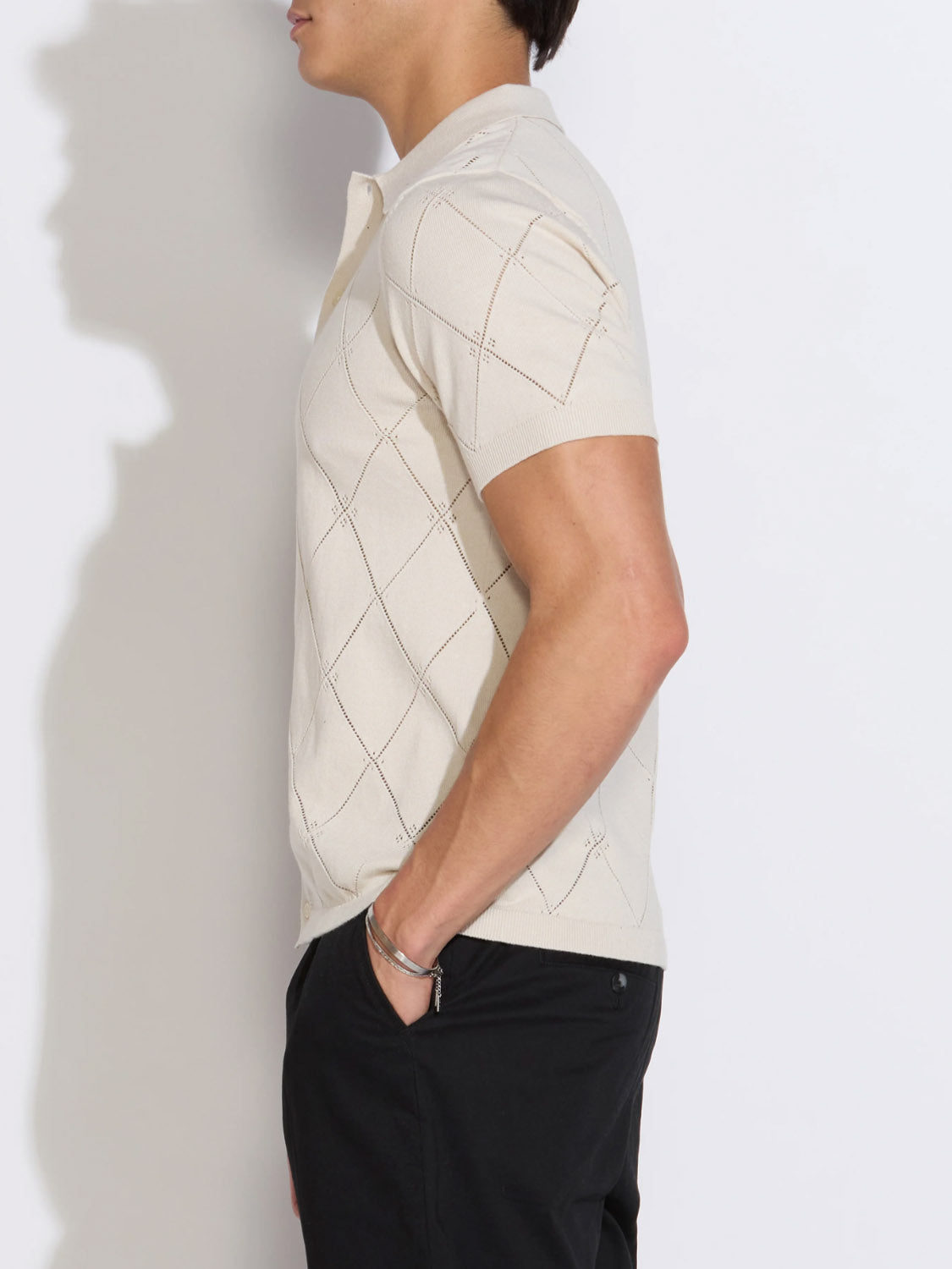 A person stands in profile against a white background, wearing Civil Society’s Aleppo Full Placket Sweater Polo with a subtle diamond pattern and black pants, one hand in their pocket.