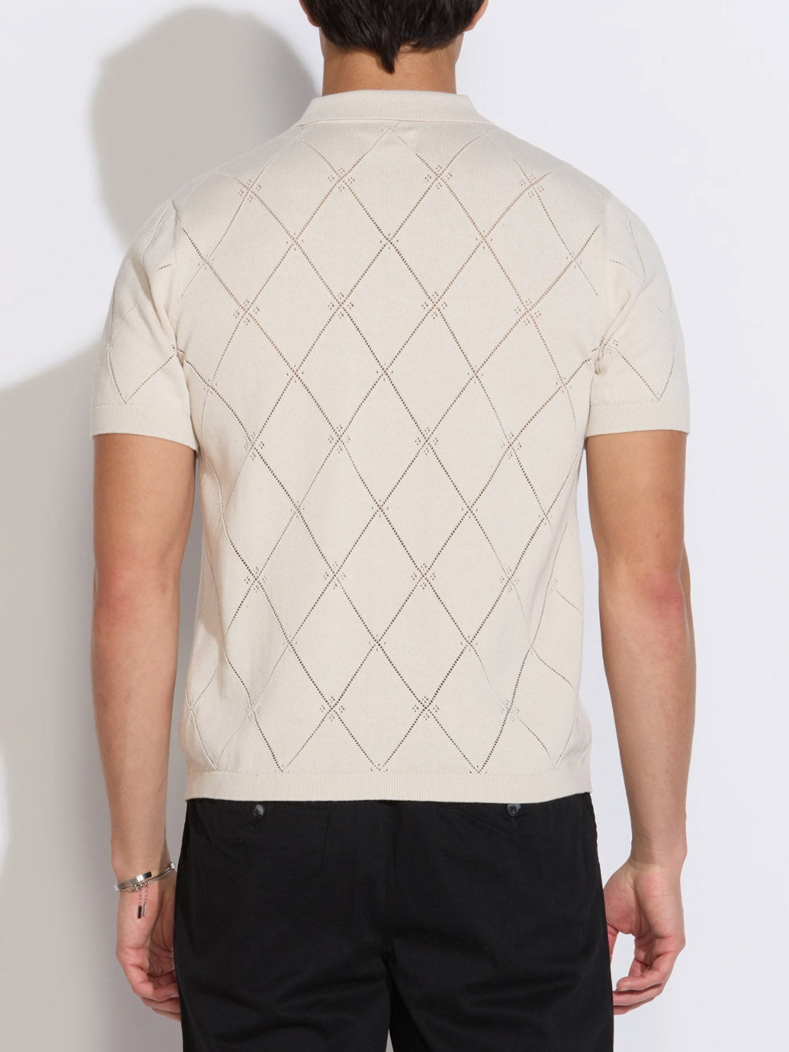 A person is shown from the back wearing the Civil Society Aleppo Full Placket Sweater Polo in cream with a diamond pattern and short sleeves, paired with black pants, against a plain white background.
