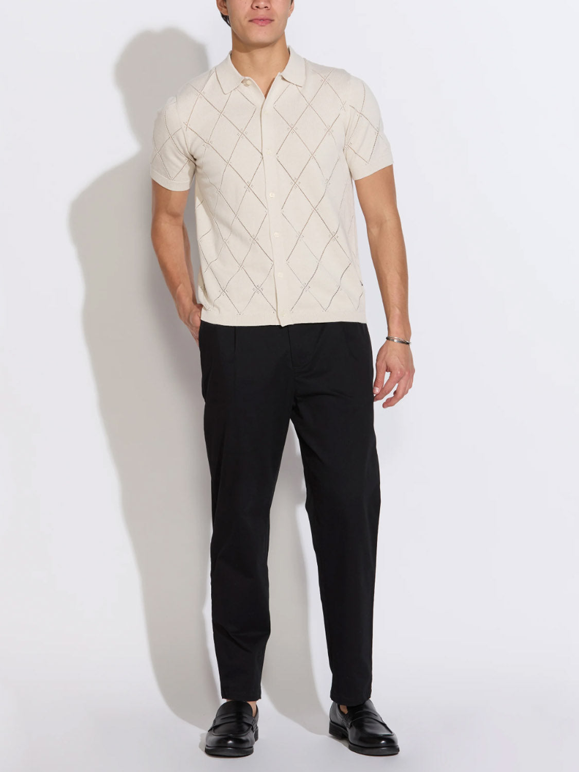 A man stands against a white background wearing the Civil Society Aleppo Full Placket Sweater Polo, a cream short-sleeve with a subtle diamond pattern, paired with black trousers and loafers. His arms are relaxed at his sides.