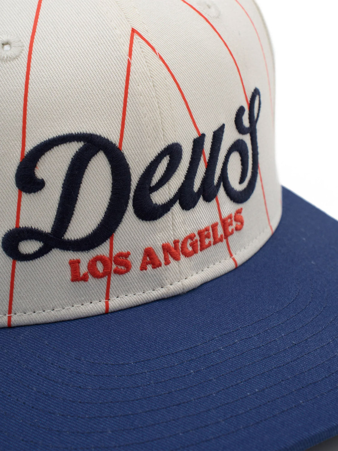 Close-up of the Alvia Cap by Deus, a cream and blue vintage-style cotton twill baseball hat with red pinstripes. Features "Deus" embroidered in black script and "Los Angeles" in red capital letters underneath.