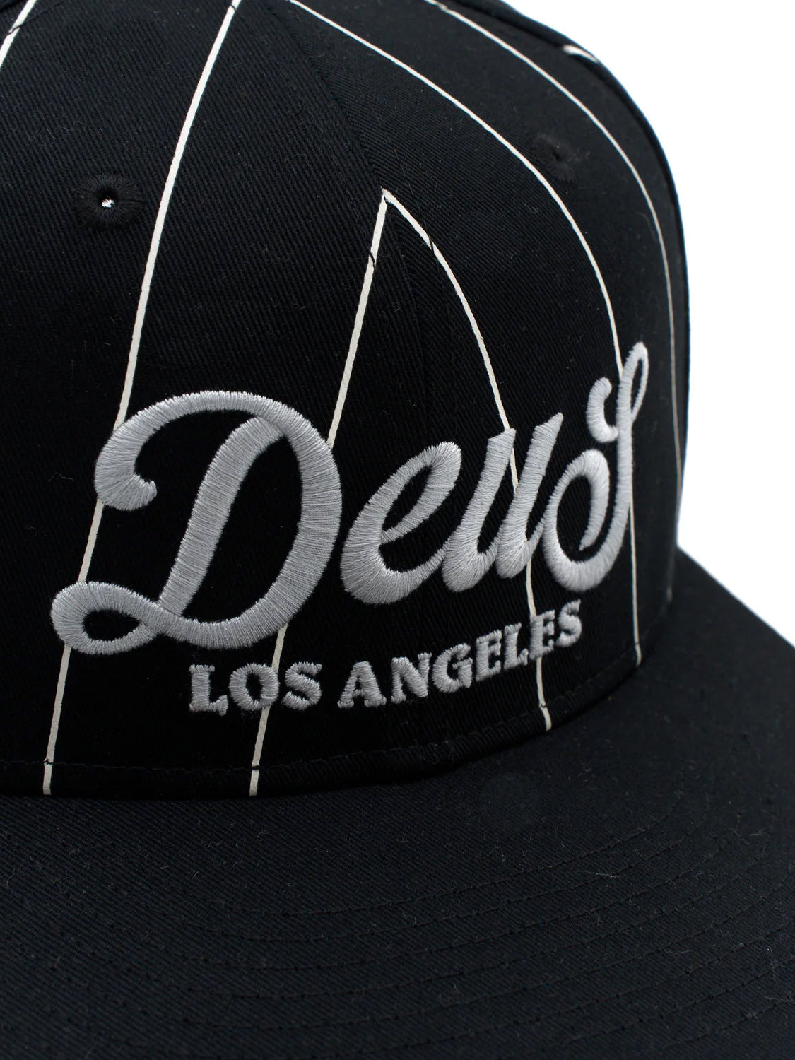 The Deus Alvia Cap is a vintage-style black cotton twill hat with white pinstripes, raised "Deus LOS ANGELES" embroidery on the front, and a classic snapback closure.