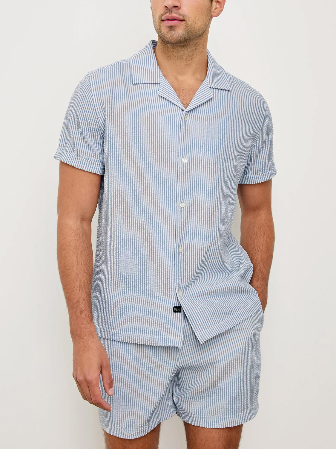 A man wears the Rails Amalfi Seersucker Shirt, featuring light blue and white stripes, with matching shorts. He stands with one hand in his pocket against a plain off-white background.