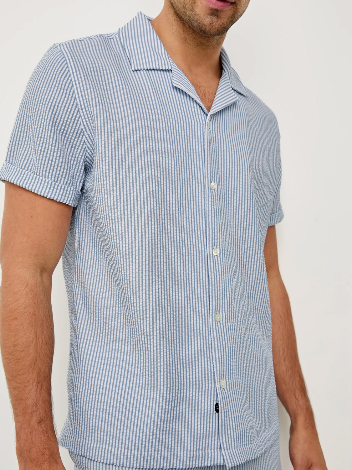 A man wears the Amalfi Seersucker Shirt by Rails, featuring light blue and white vertical stripes, short sleeves, a relaxed fit, and a collared design. He stands against a plain white background with his head partially out of frame.