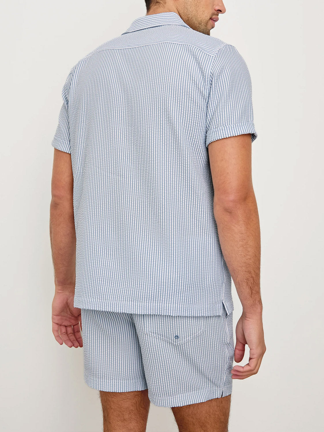 A man is seen from behind wearing the Rails Amalfi Seersucker Shirt in light blue and white stripes, paired with matching shorts. The outfit features a relaxed fit for a casual, summery look.