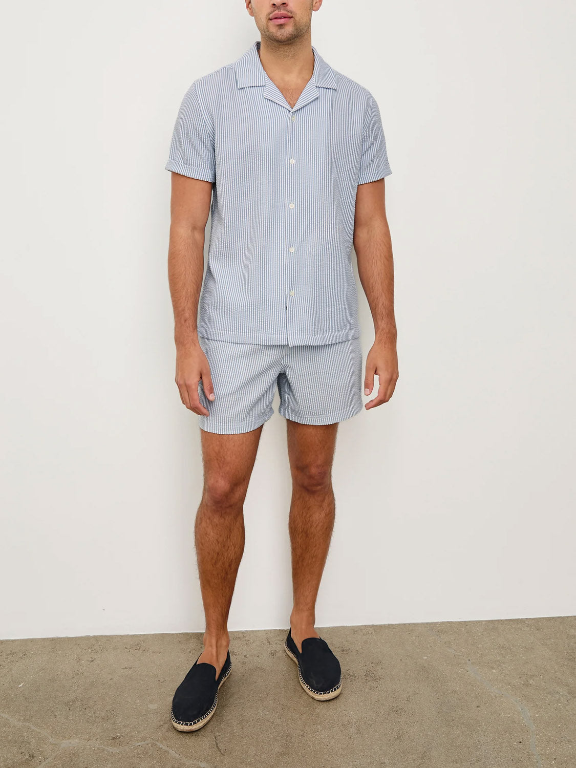 A man stands against a plain white wall wearing the Rails Amalfi Seersucker Shirt in light blue and white pinstripes, paired with matching shorts and black espadrilles.