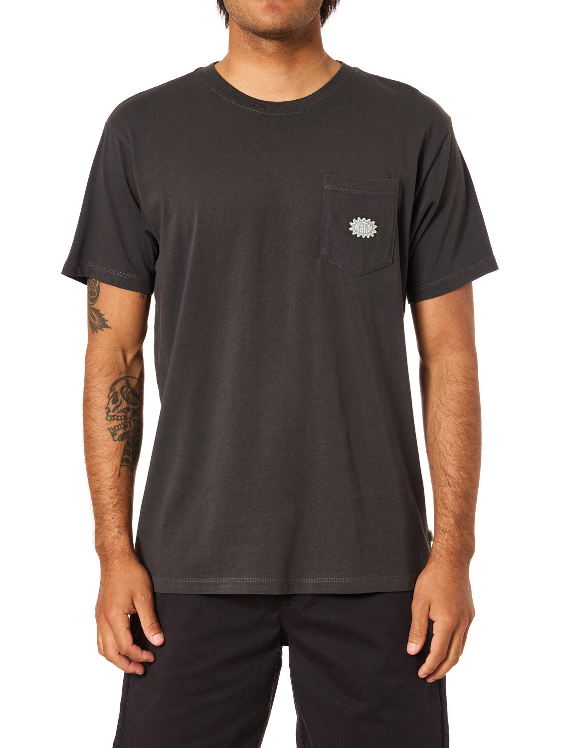 A person wearing a Katin Amoeba Pocket Tee by Katin, made from organic cotton, faces forward. The plain black shirt features a small white custom typography graphic on the chest pocket. They have visible tattoos and wear black shorts.
