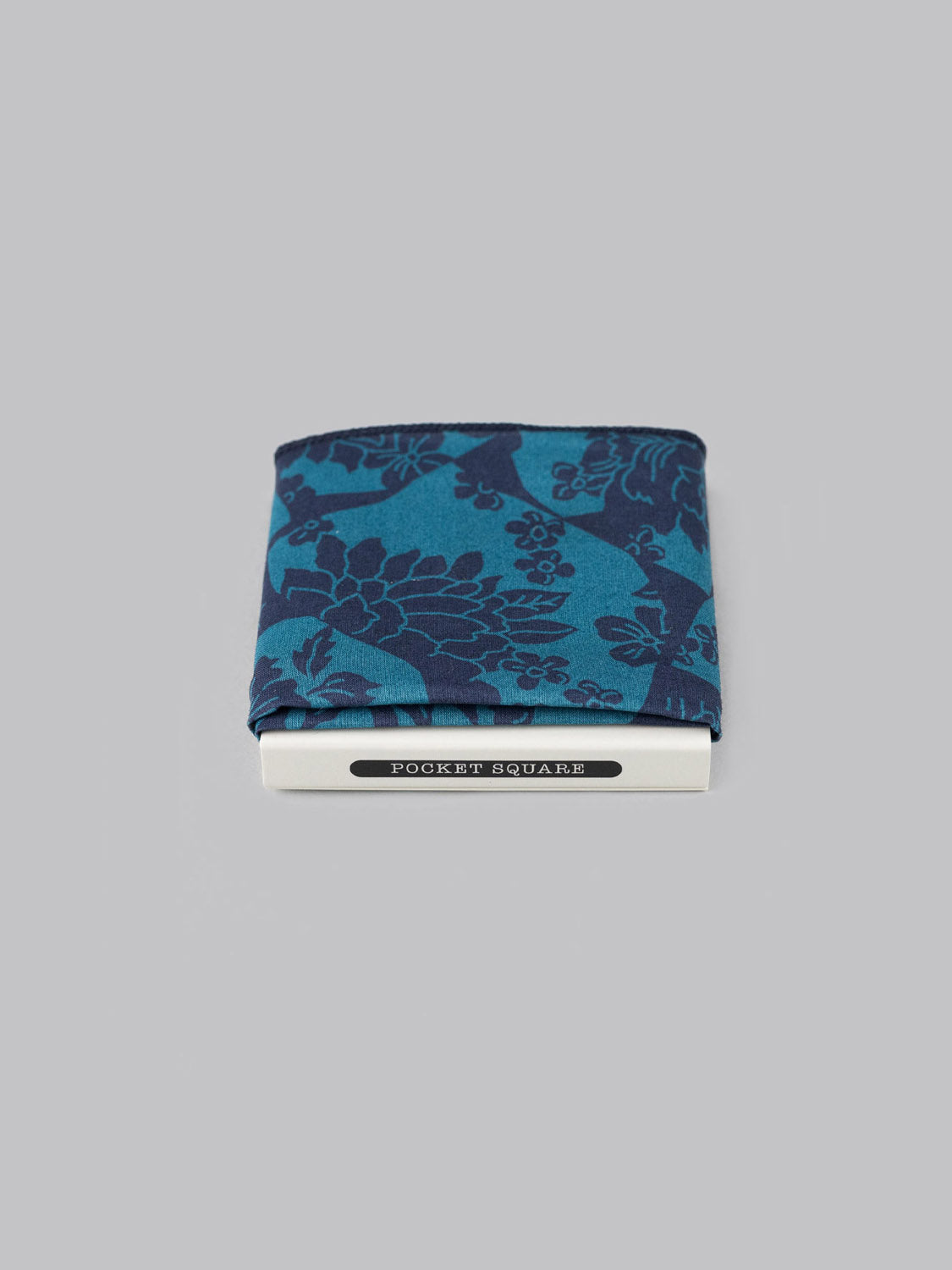 A teal and navy Liberty™ Ancient Pocket Square by ID Menswear, made from soft Tana Lawn cotton, is neatly folded on a plain grey background with a visible POCKET SQUARE label beneath.