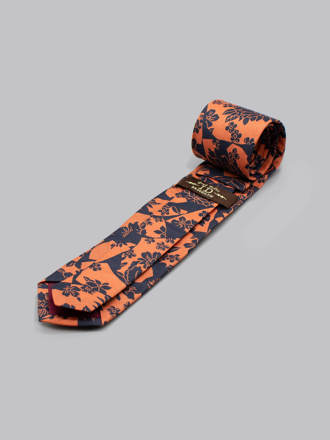 The Liberty of London Tana Lawn™ Ancient Tie by ID Menswear—a rolled-up orange tie with a dark blue floral pattern—lies on a light gray background, showing its label inside. Handmade in Brooklyn from soft Tana Lawn cotton.