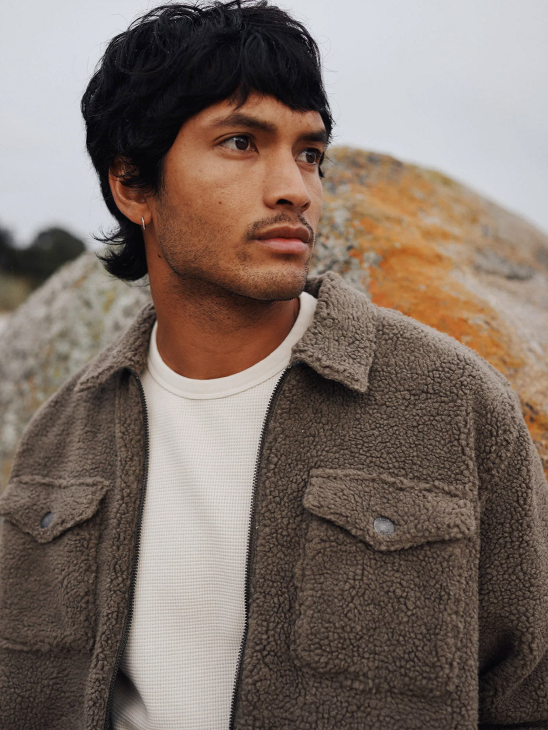 A man with dark hair and facial hair wears the Katin Anderson Sherpa Jacket over a light shirt, standing outdoors in front of a large rock and gazing thoughtfully into the distance.
