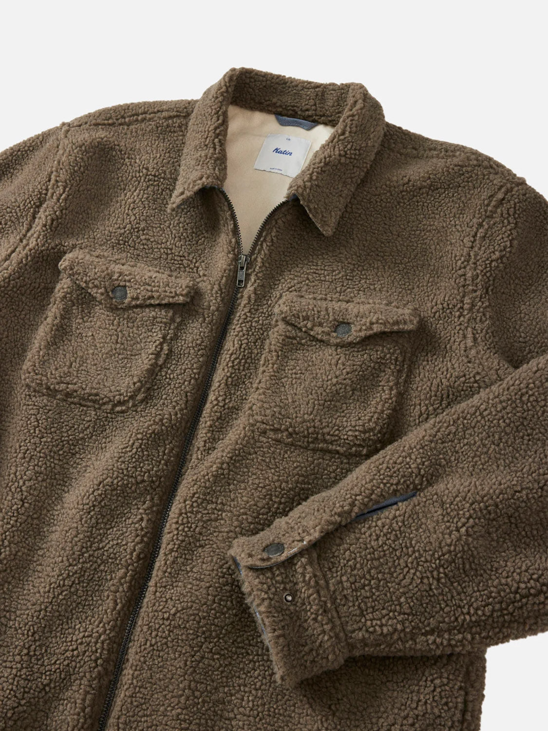 The Anderson Sherpa Jacket by Katin is a brown, zip-up polyester sherpa jacket with two buttoned chest pockets, a collar, a visible brand tag inside the neck, and a cozy fleece texture. The left sleeve is partially folded.