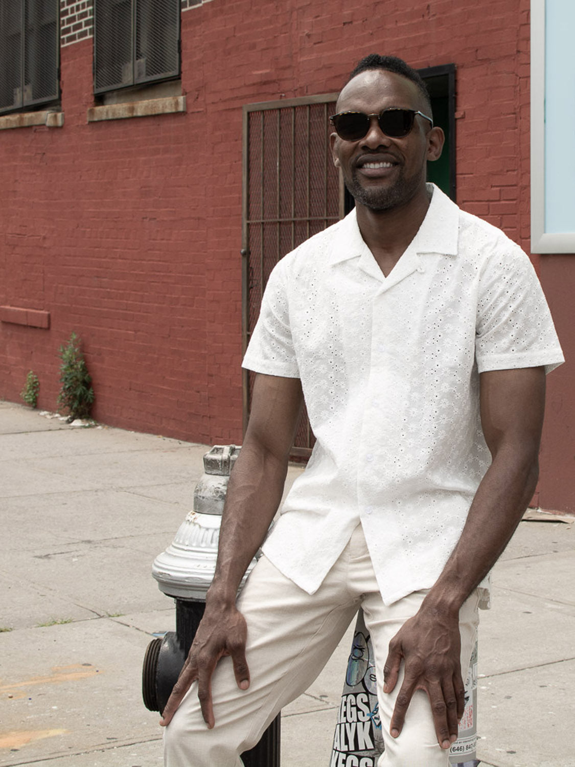 A man in an Anglaise Embroidery Shirt by ID Menswear and sunglasses sits on a fire hydrant on a city sidewalk, smiling, with a red brick building in the background.