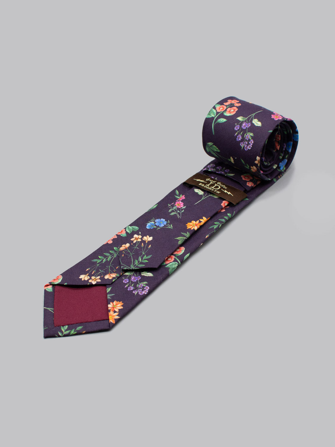 Handmade in Brooklyn, the ID Menswear Liberty of London Tana Lawn™ Annie tie features a dark purple floral pattern, maroon back tip, and visible brand label. Shown rolled up on a light gray background.
