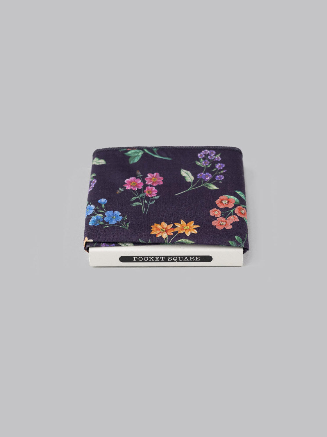 The Liberty™ Annie Pocket Square by ID Menswear, made from Tana Lawn cotton with a vibrant floral print on a dark background, is folded and partially placed in a white POCKET SQUARES box against a light gray backdrop.