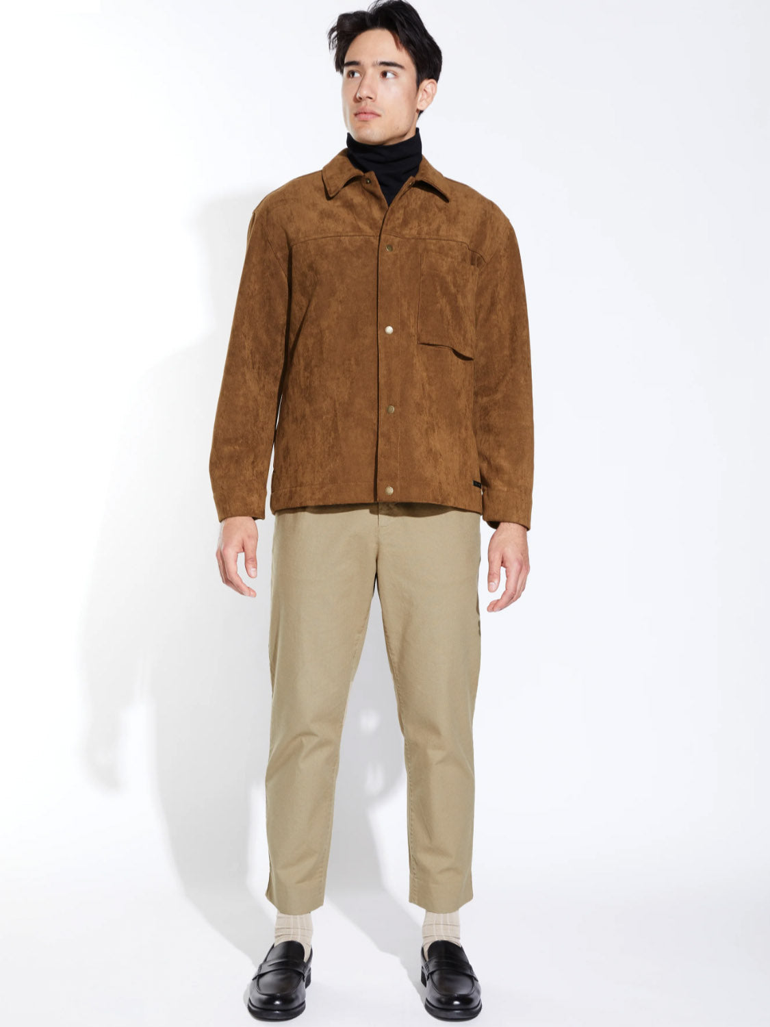 A man stands against a white background wearing the Civil Society Ansel Boxy Fit Suede Shirt Jacket in brown, paired with a black turtleneck, tan pants, white socks, and black loafers. He looks slightly to the side with arms relaxed at his sides.