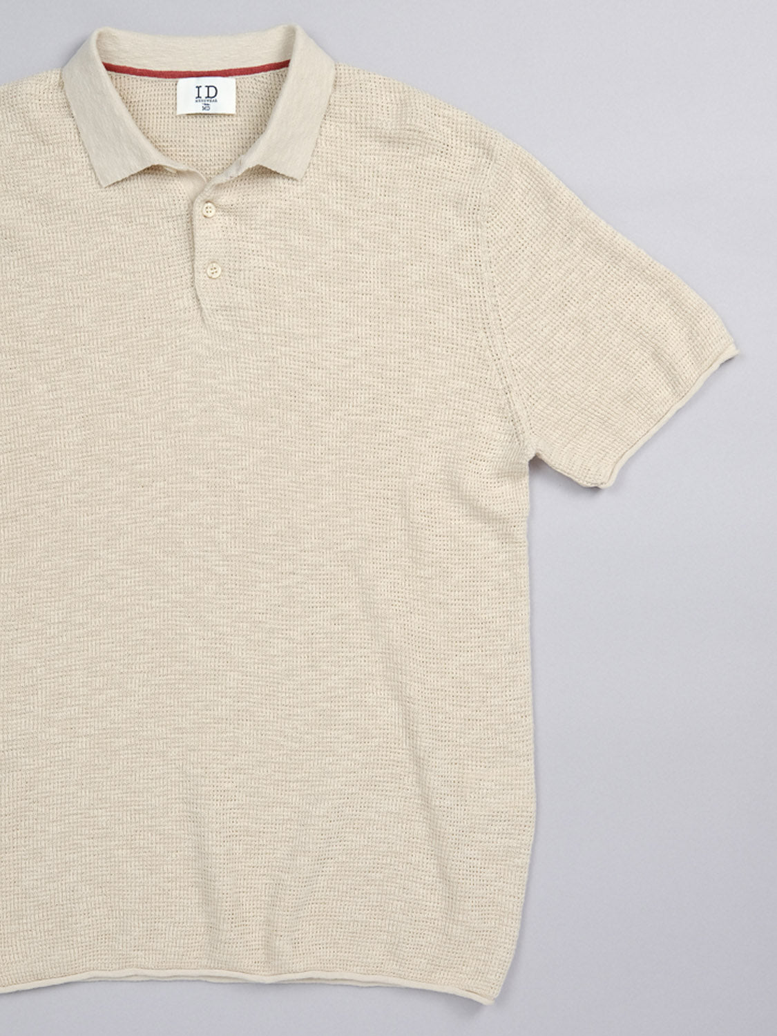 The Athens Waffle Knit Polo by ID Menswear is a beige, short-sleeve shirt with a textured waffle-knit design and button-up collar. Laid flat on a gray background, it features a subtle inside collar label, ideal for casual style.