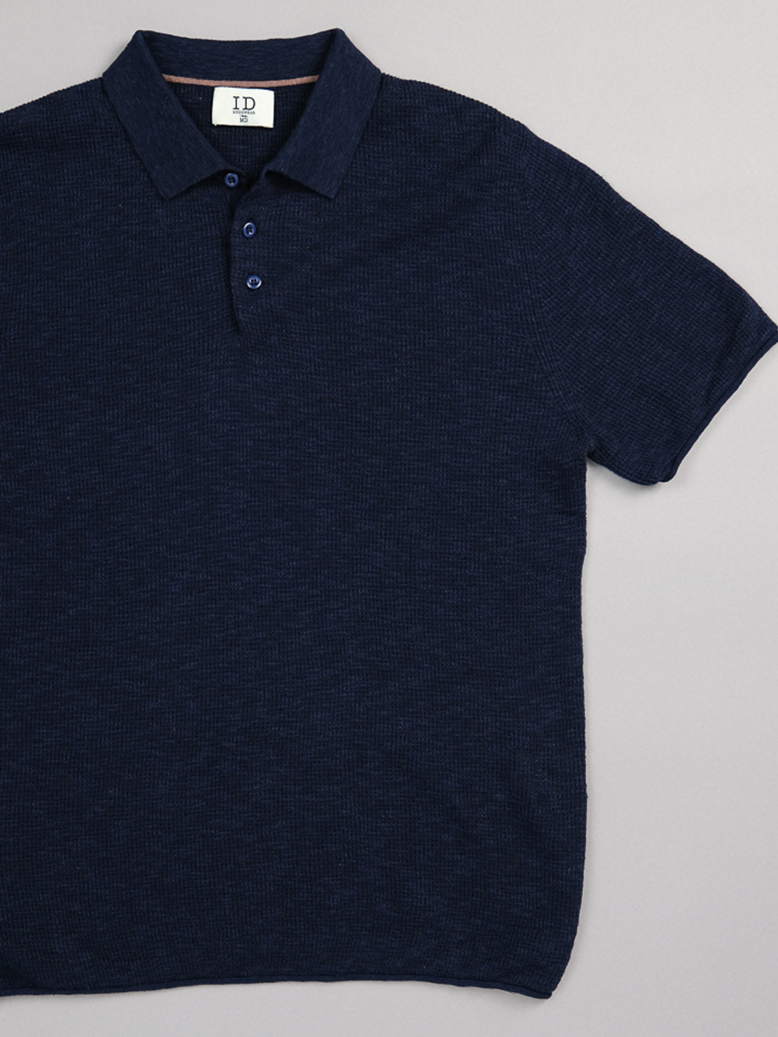 The Athens Waffle Knit Polo by ID Menswear, a smart-casual navy blue shirt with a collared neckline, is laid flat on a light gray surface. It showcases three dark buttons and has an interior collar tag displaying the initials ID.