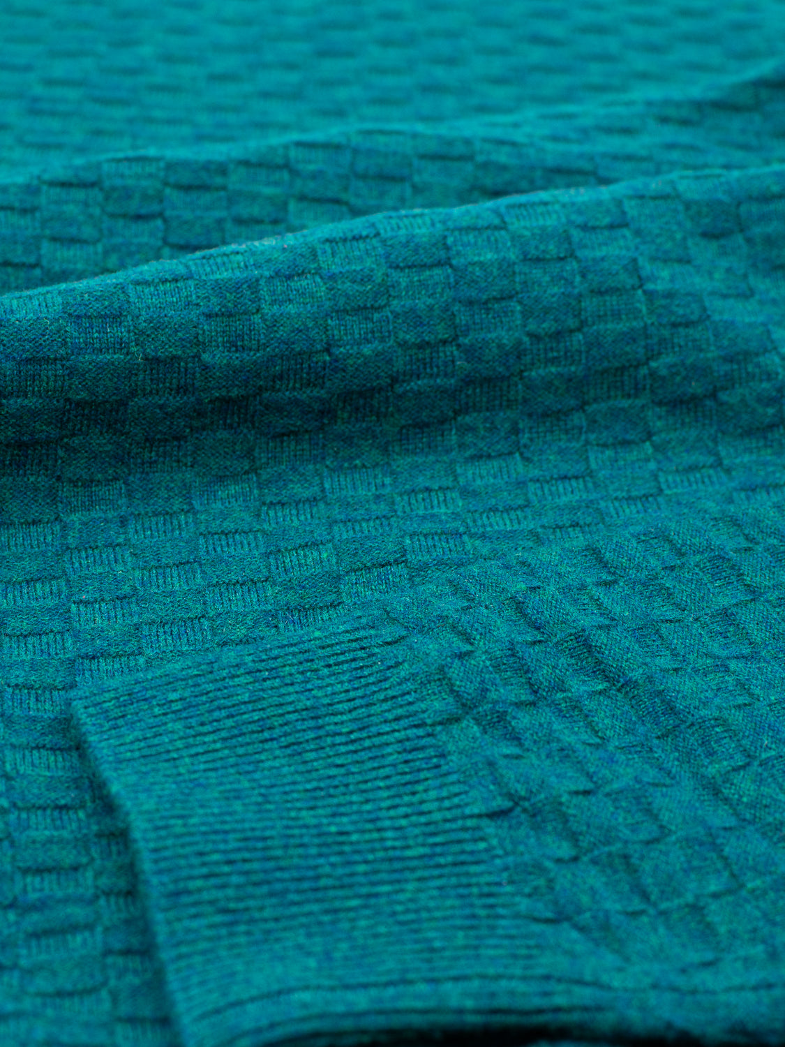 Close-up of the Stockholm Merino Wool Sweater by ID Menswear in teal green, showcasing a textured square checker pattern. This classic crewneck features soft, lightweight knitted fabric with a folded sleeve in view and thin ribbed cuffs.