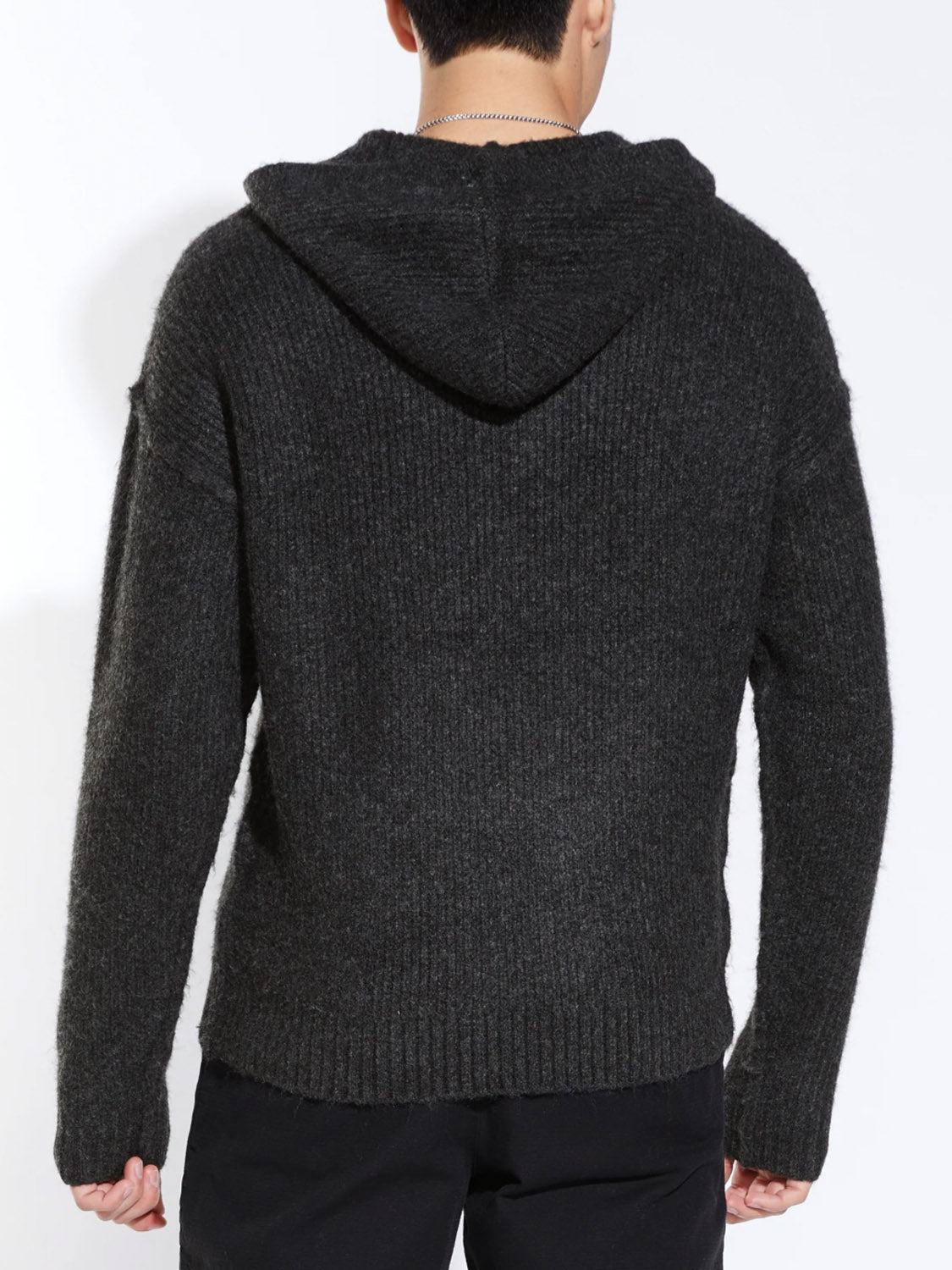 A person stands with their back to the camera, wearing the Civil Society Avery Oversized Sweater Hoodie in dark gray and black pants. The hood is down, showing short dark hair and a silver necklace.