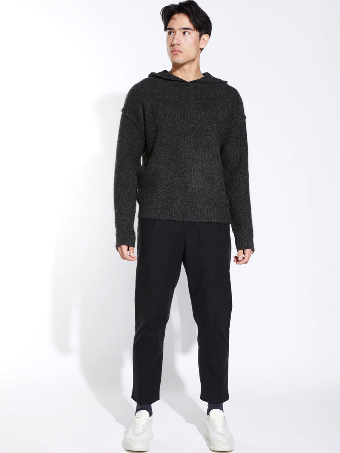 A man faces slightly to the side, wearing the Civil Society Avery Oversized Sweater Hoodie in dark gray, paired with black pants and white sneakers, against a plain white background.